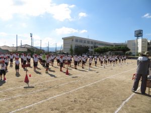 晴天の下、運動場に児童の皆さんが等間隔に間をあけて整列し、運動会全体練習に参加している様子を写した写真
