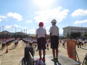 晴天の下、運動場には児童の皆さんが等間隔に間をあけて整列しており、手前に置かれている朝礼台に代表の児童2名が立っている運動会全体練習の様子の写真