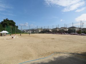 運動場のコーナーを 走っている児童、運動場の奥には児童の皆さんが集まっている様子が分かる全体練習風景の写真