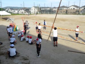 運動場の一角に数名の先生方と児童の皆さんが集まり、運動会で使われる、玉入れのかご、三角コーンなどが置かれた場所で、長い棒の両端を生徒が2人で持つなど係活動の準備をしている様子の写真