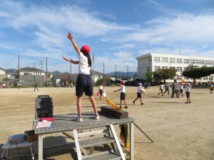 運動場に置かれた朝礼台に赤色の帽子をかぶった女子児童が立ち、両手を斜め上に挙げ、体操をしており、運動場にも多くの生徒たちがそれぞれの活動をしている様子の写真
