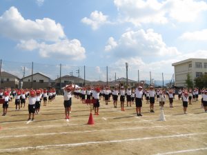 青空の下、運動場に児童の皆さんが整列して等間隔に間をあけて並び、両手を頭の上で伸ばし、準備運動をしている様子の写真