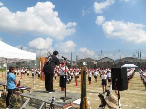 青空の下、運動場に整列している児童の皆さんが見守る中、児童代表の生徒が朝礼台に上がった関係者の方から優勝旗を受け取っている様子の写真