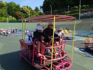 大きな自転車のような乗り物で、後ろには数名の生徒が座っており、先頭に座る3名の生徒でペダルを漕いで前に進んでいる様子の写真