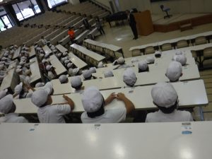 教壇を中心に、座席が後方になるほど階段式に高くなっている教室で、帽子を被った生徒たちが正面に立つ先生の話を聞いている様子の写真