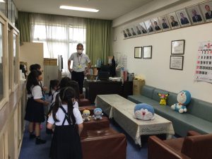 壁に多くの顔写真が飾られた校長室のような場所で、応接ソファーの近くに立って室内を見学をしている生徒たちの写真