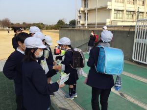 校舎近くの場所で、子どもたちが募金箱のようなものを手に持って並んでおり、ランドセル姿の子どもが募金をしている様子の写真