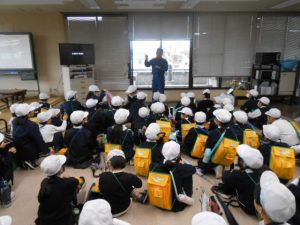 屋内で一人の消防署の関係者が子どもたちに話をしており、子ども達は座って興味深そうに話しを聞いている様子を後方から写した写真