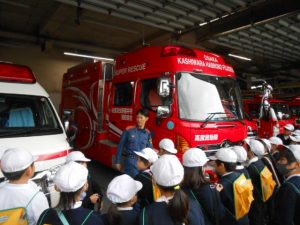 救急車、消防車などの救急車両が数台停まっている建物内で、児童の皆さんが消防署の男性職員の話を聞いている様子の写真