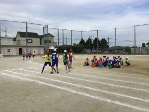 赤、黄色、緑、青色のゼッケンをつけ、4名の児童がコーナーを走っており、中継地点で4名の児童が待ち構えているリレーの練習風景の写真
