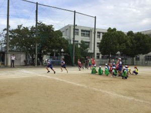 運動場のフィールド内でチームごとに分かれて座って待機している児童とトラックの自分のコースを足を高く上げて走っている児童が写っている運動会練習風景の写真