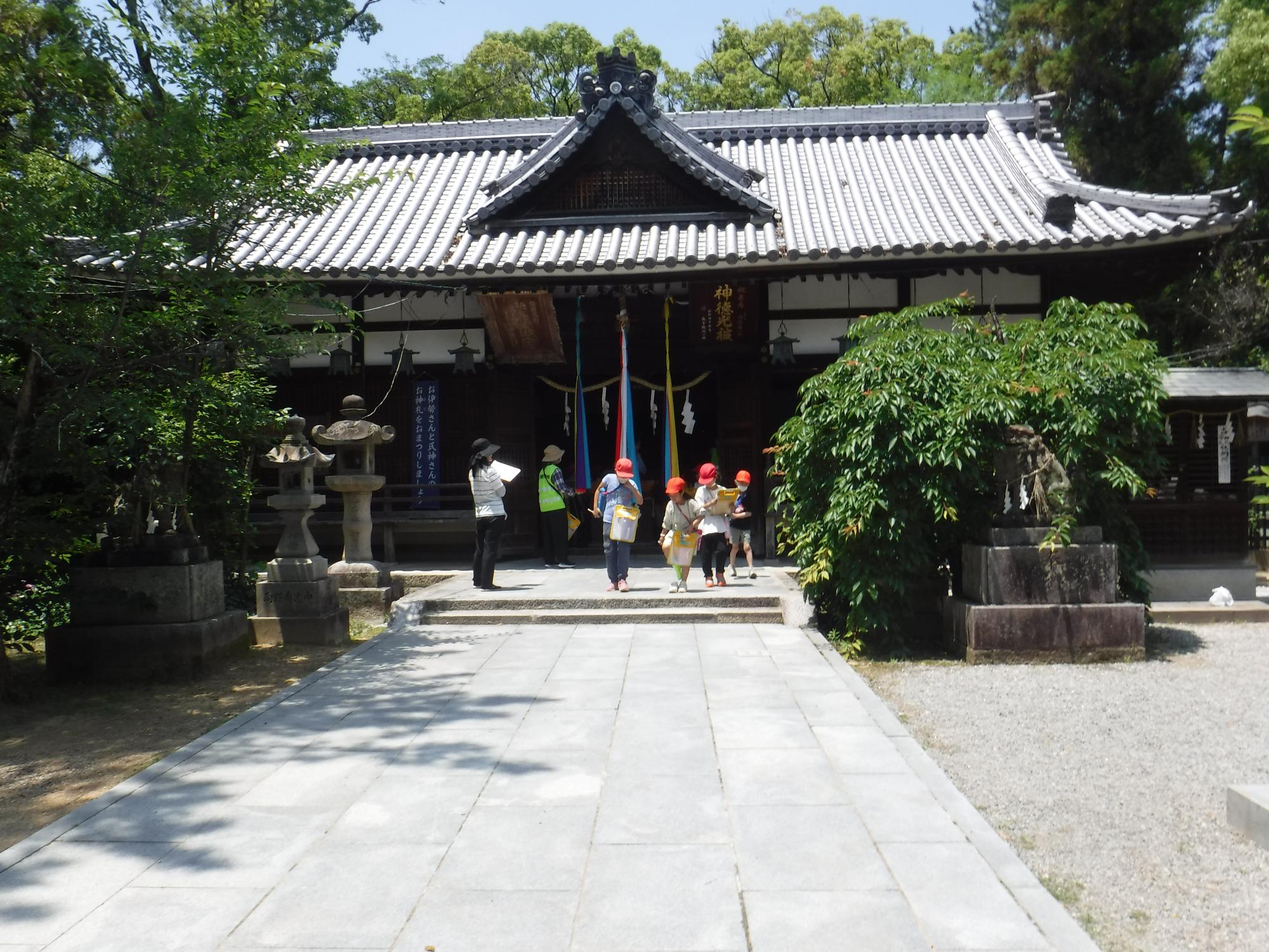 木々に囲まれた神社の拝殿の前で、赤白帽子を被った数名の児童と保護者の方が歩いている写真