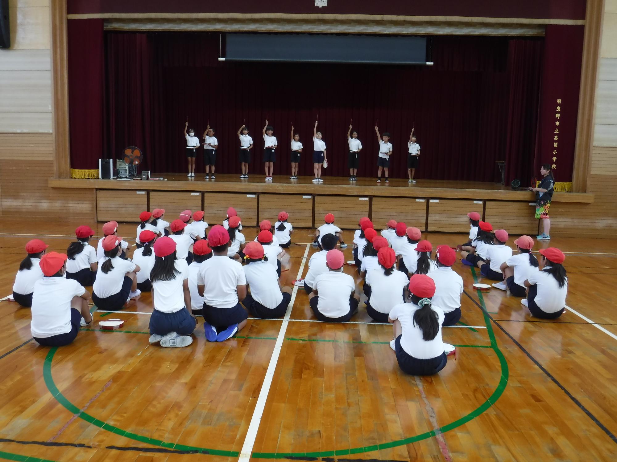 体育館のステージ上に横一列に並んだ体操着姿の9名の実行委員の児童が、右手を挙げてエイサーを踊っている様子を他の児童たちが見ている写真