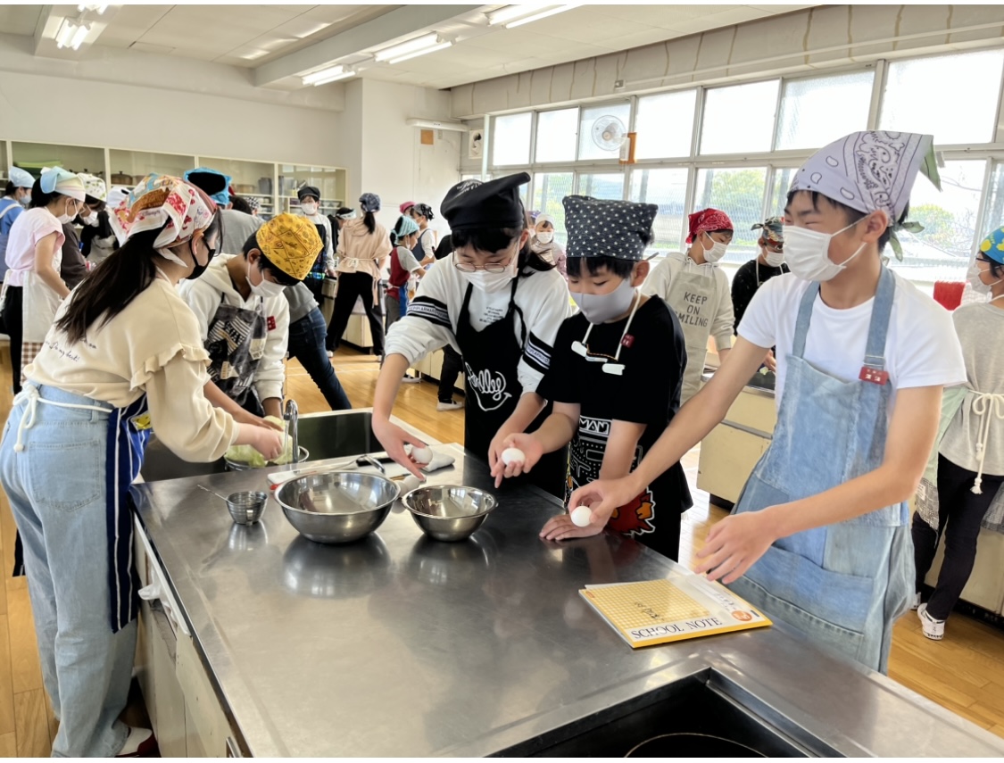 三角巾やエプロンを着用した児童たちがグループごとに分かれ、2名の児童が野菜を洗い、3名の児童がボウルに卵を割ろうとしている調理実習の様子の写真