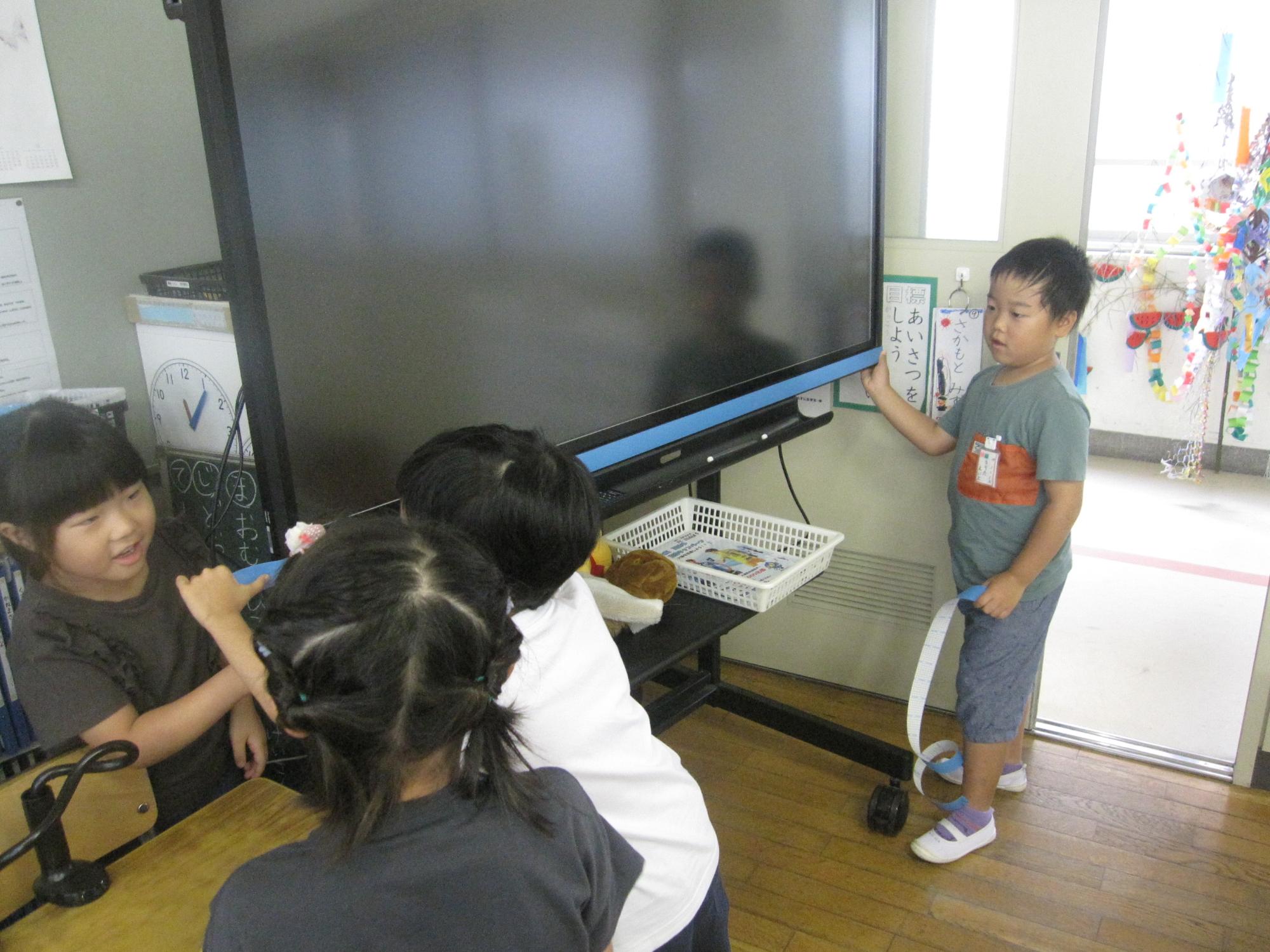 教室の入り口近くに設置された大画面のテレビモニターの横位置に児童たちが水色の紙テープを置き、長さを測っている様子の写真
