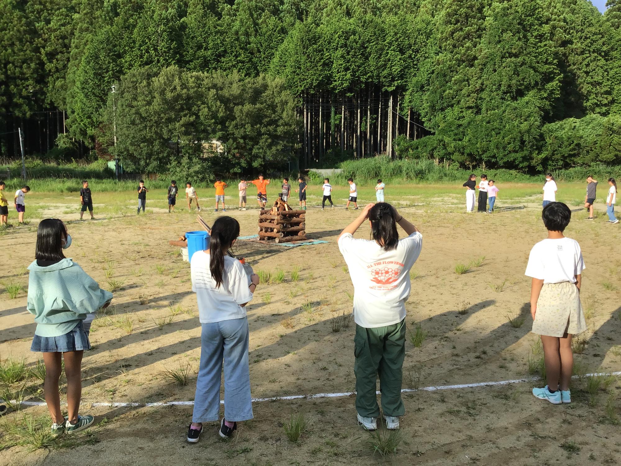 沢山の木々に囲まれた広場の中央に木材が積まれ、周りを児童たちが囲んでいるキャンプファイヤーの様子の写真