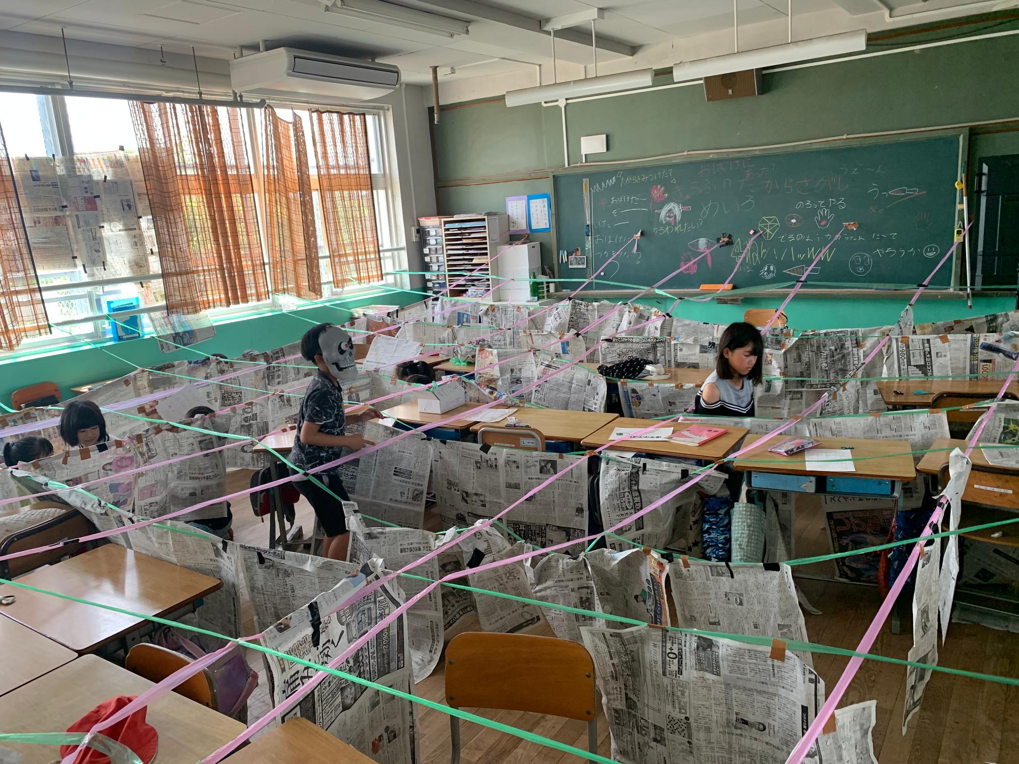 机や新聞紙で壁を作り、教室内をピンクや緑のロープで交差して区切っている中央でお面を被った男の子が立ち女の子を見ている出し物の写真