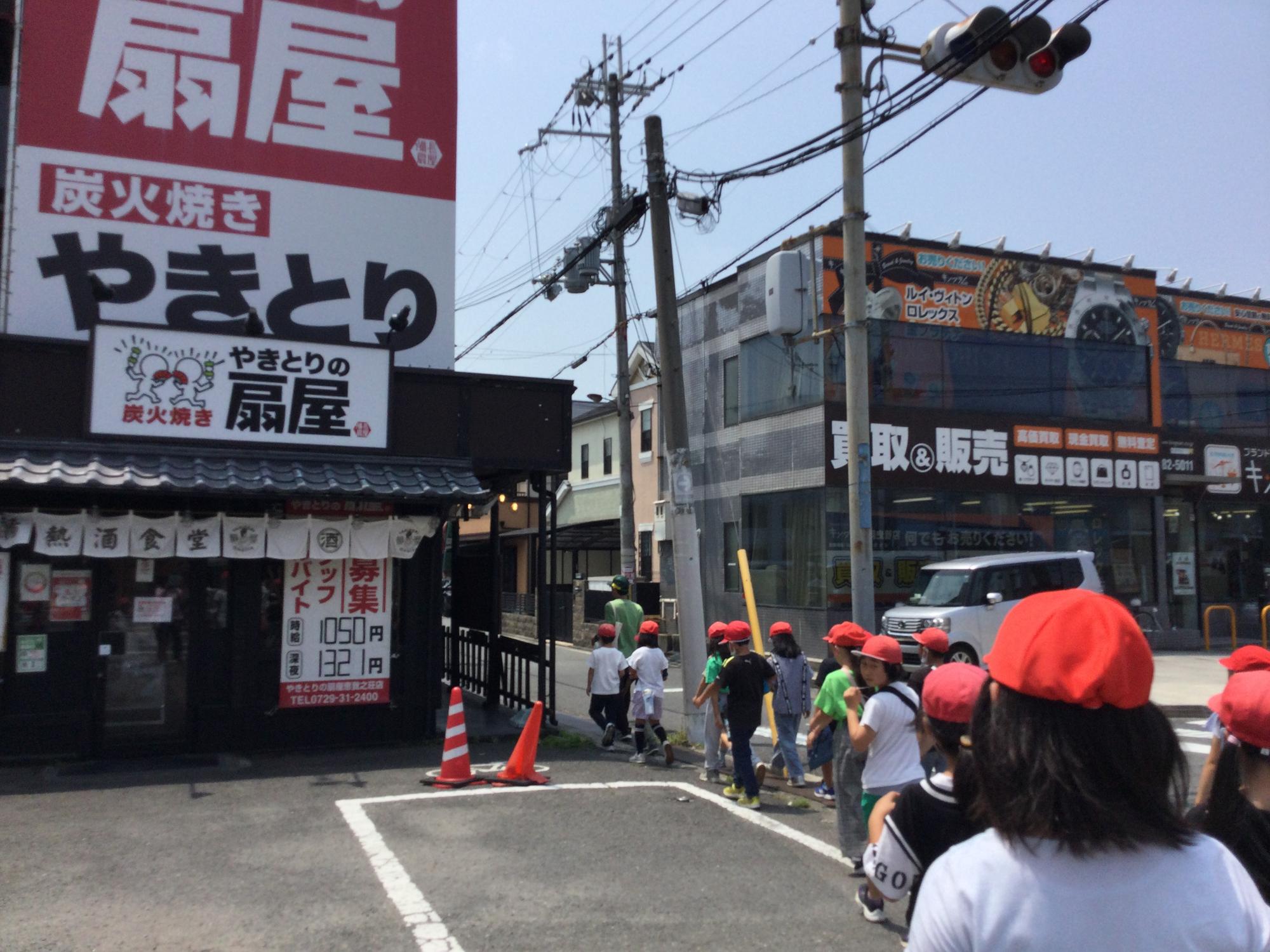 居酒屋や買取の店舗の間の細い道路を、先生を先頭に赤白帽子を被った児童たちが2列に並んで歩いている写真