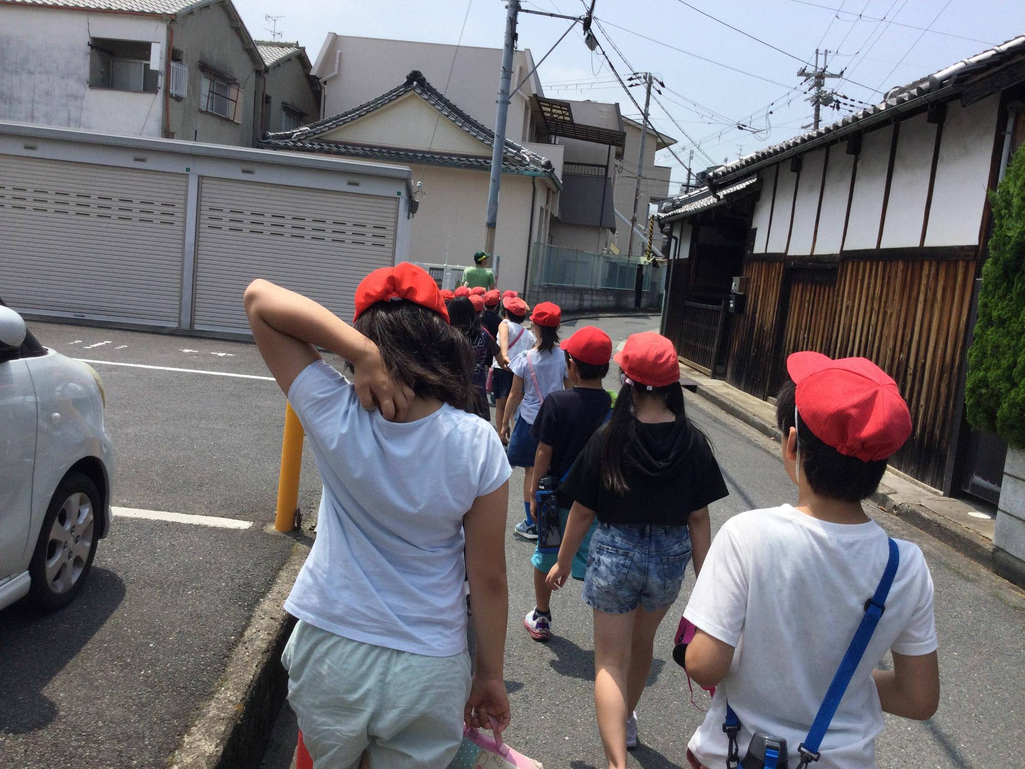 趣のある建物と駐車場の間の細い道路を、先生を先頭に赤白帽子を被った児童たちが2列に並んで歩いている後ろ姿の写真