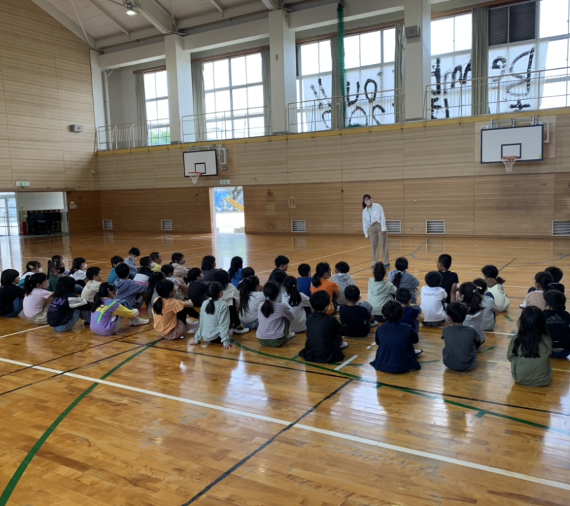 体育館内の床に児童たちが4列に体育座りして並び、向かい合って立っている女性教師の話を聞いている後ろ姿の写真