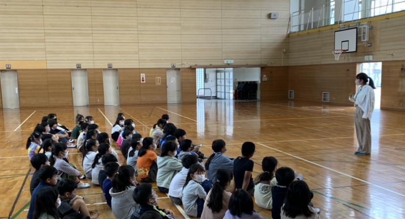体育館内の床に4列に体育座りして並んだ児童たちが、向かい合って立っている女性教師の話を聞いている様子を横から撮影した写真